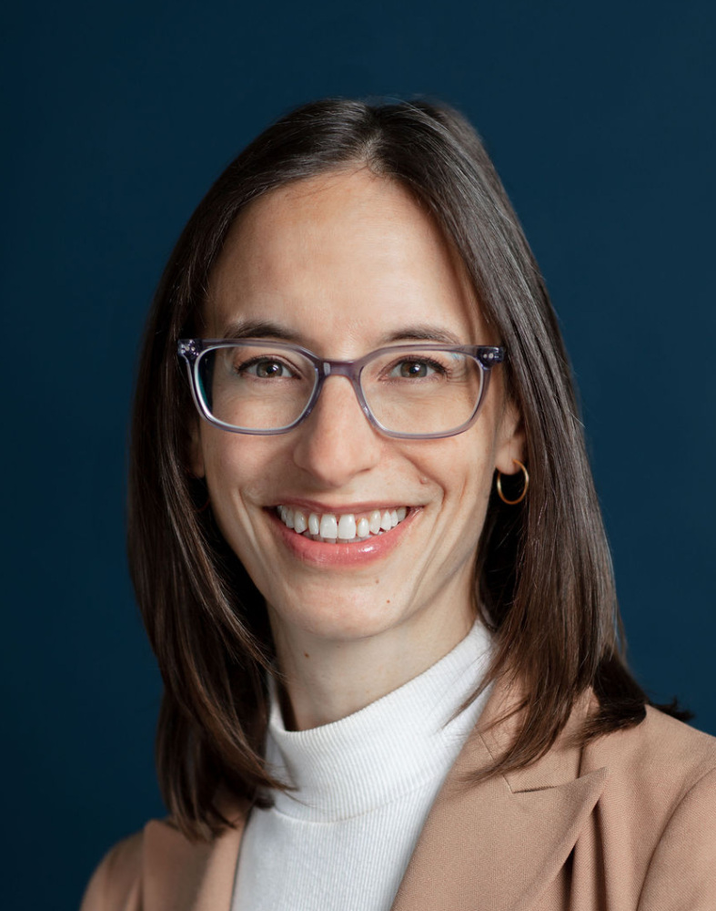Woman with straight brown hair, wearing glasses, a beige blazer, and a white turtleneck, smiling in front of a dark blue background.
